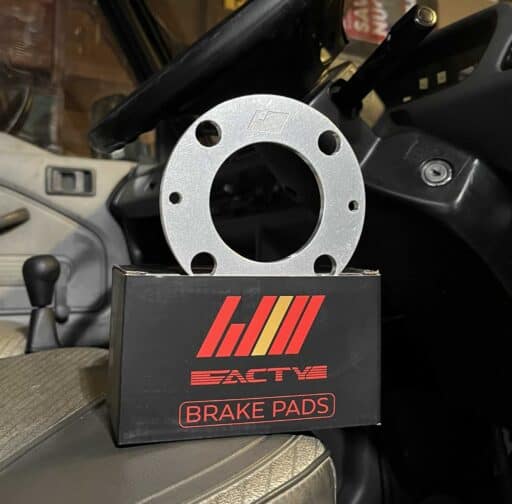 Aluminum brake caliper spacer disc on top of black packaging box with "EACTIV Brake Pads" logo, inside a vehicle; aftermarket performance brake part for enhanced vehicle stopping power.